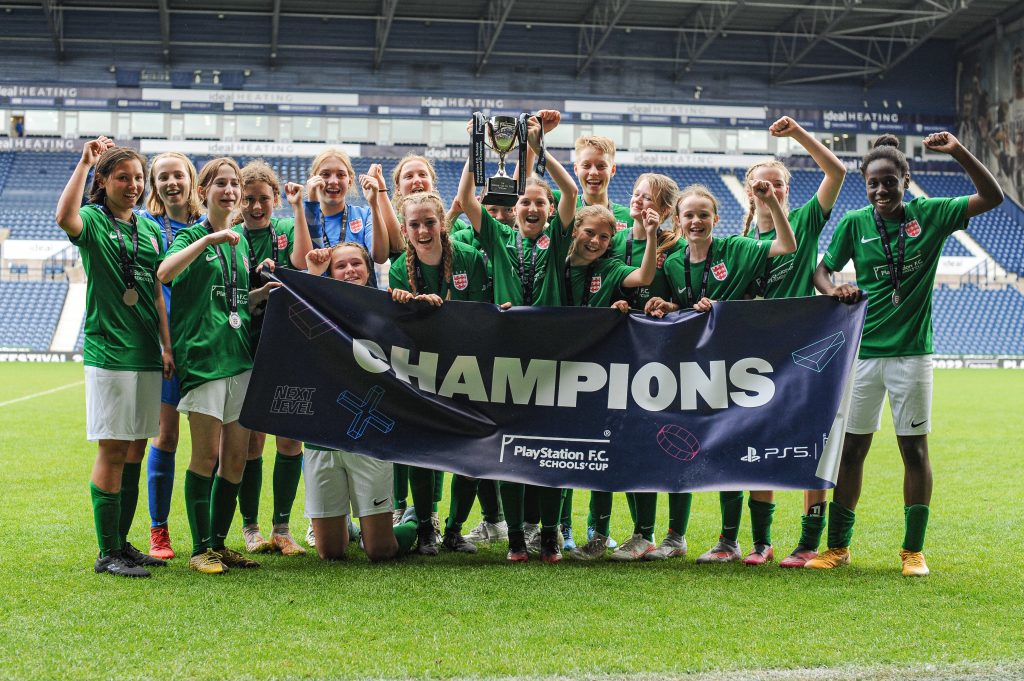 Surbiton High School U14 Girls' Cup champions holding a winners' banner on the pitch at West Bromwich Albion's The Hawthorns Stadium