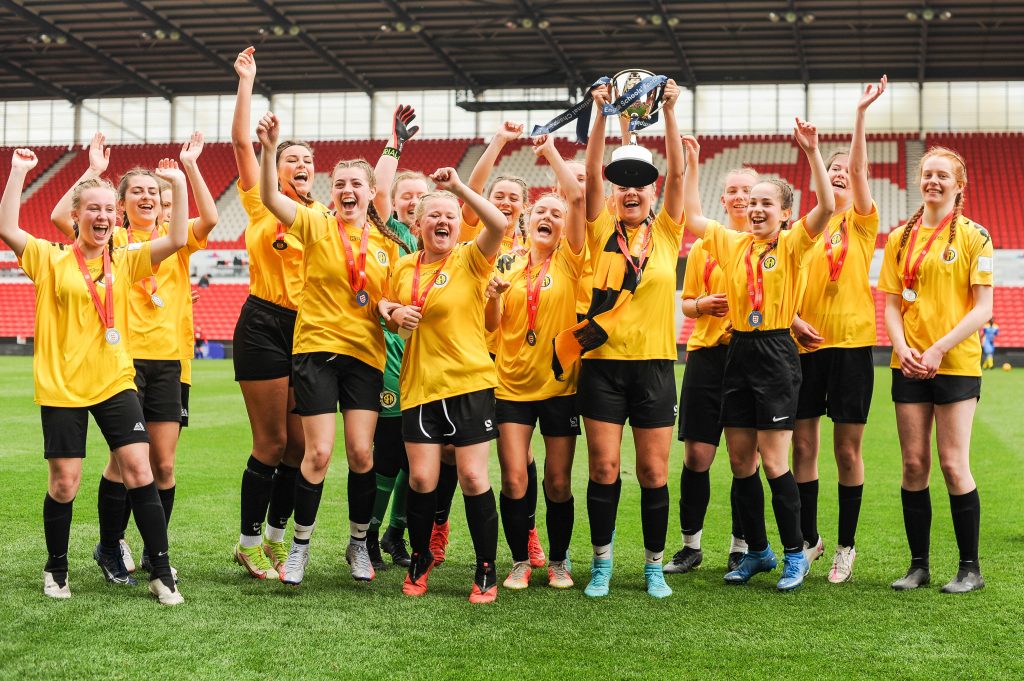 Shenfield High Schools' U16 Girls' team in their yellow and black kit, lifting the trophy and celebrating as they win their National Cup title at Stoke City FC